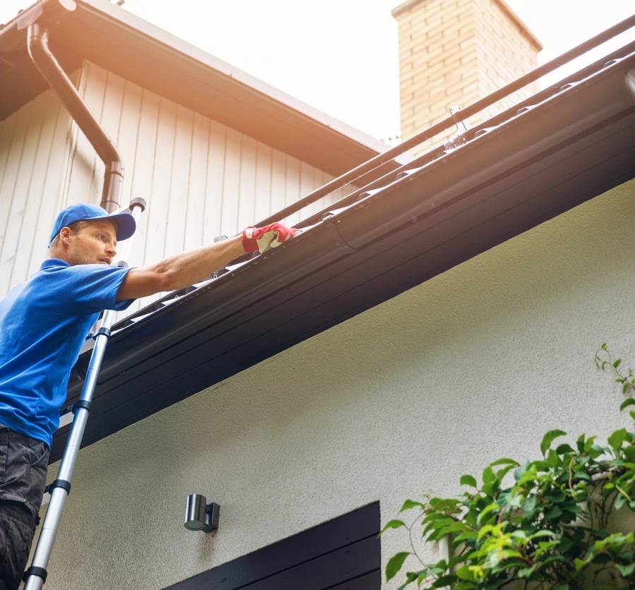 man on ladder cleaning house gutter from leaves and dirt